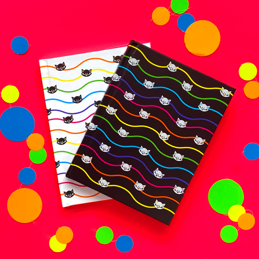 Two journals on top of a bright red surface. Top journal is black, with wavy rainbow pinstripes and white cats. Bottom journal is white with rainbow pinstripes and black  cats. 